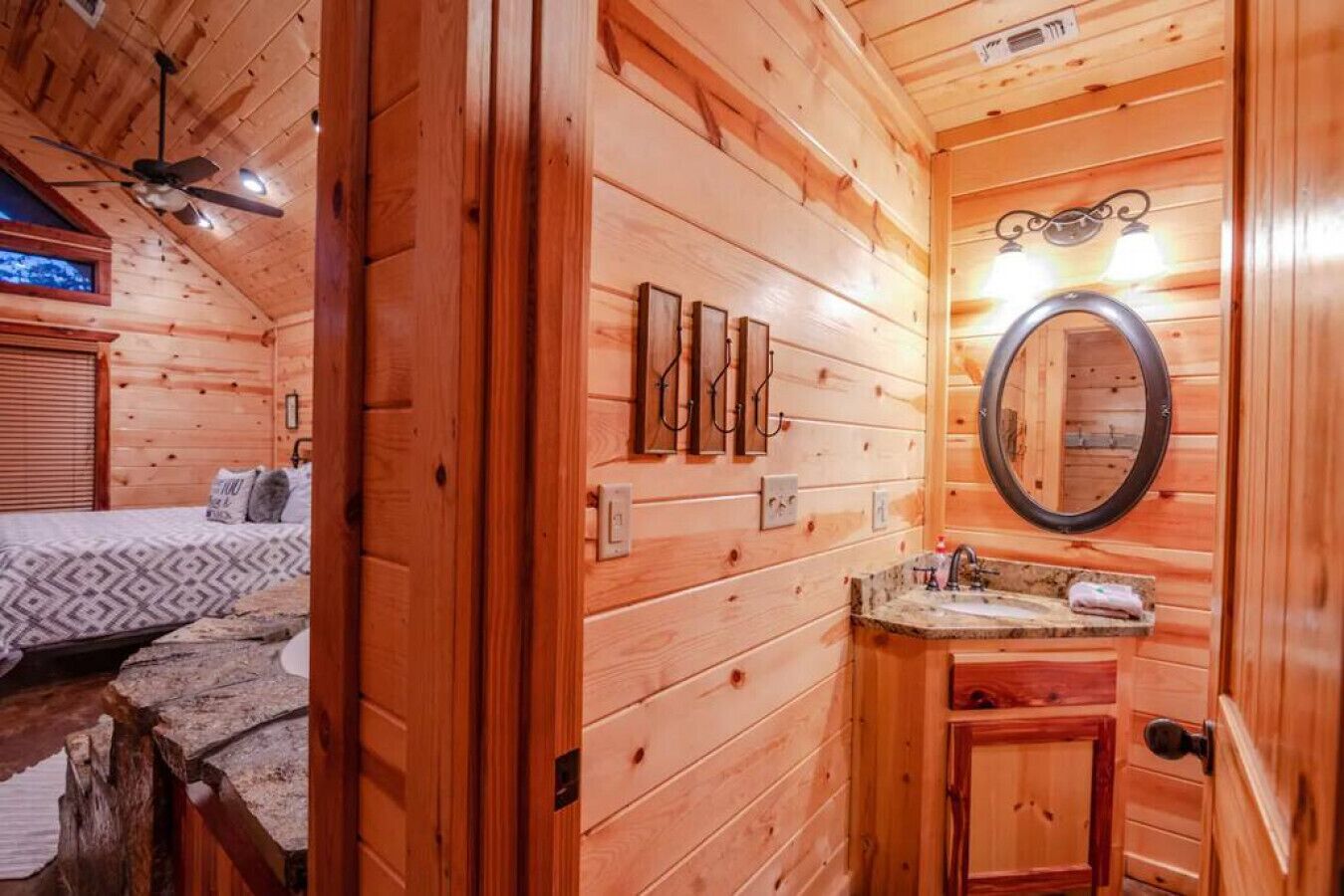 Ein Interieur einer Holzhütte mit einem Bett, einem Badezimmer mit Spiegel und Waschbecken sowie einem Deckenventilator.