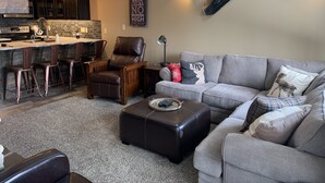 A modern living room with a grey sofa, a leather chair, and a coffee table. There is a kitchen area with a bar and stools. - Schweitzer true ski in ski out condo (Sandpoint)