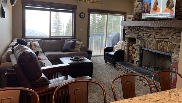 A cozy living room with a stone fireplace, a large window with a view of trees, a comfortable sofa, and a dining area. - Schweitzer true ski in ski out condo (Sandpoint)