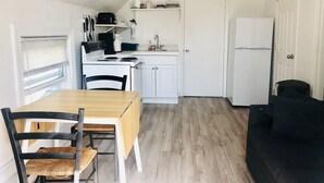 A compact kitchen with white cabinets, a stove, and a refrigerator. There is a dining table with chairs and a black sofa in the living area.