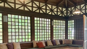 A spacious room with a high ceiling, wooden beams, and large windows. There is a long bench with cushions and a small table. - Beachfront  3-Bedroom Home in Santa Teresa with AC  (Santa Teresa, Mal Pais)