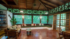 A spacious room with a wooden ceiling, large windows with green foliage outside, a sofa with cushions, a small table, and a wooden chair. - Beachfront  3-Bedroom Home in Santa Teresa with AC  (Santa Teresa, Mal Pais)