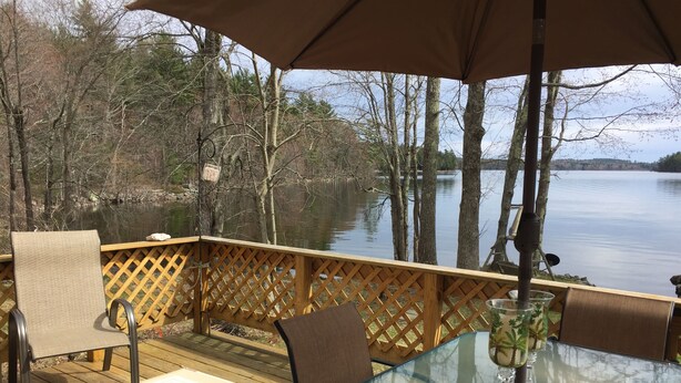 Una terraza con mesa y sillas de vidrio, con vista a un lago tranquilo con árboles y un muelle.