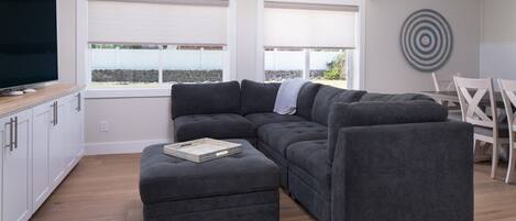 A modern living room with a sectional sofa, a small ottoman, a dining table with chairs, and a large flat-screen TV.