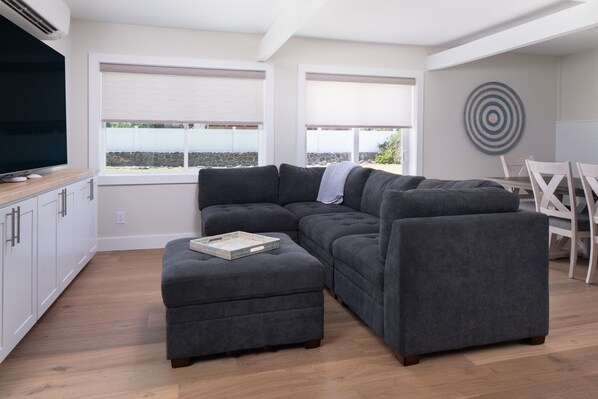 A modern living room with a sectional sofa, a small ottoman, a dining table with chairs, and a large flat-screen TV. - Charming Direct Oceanfront North Shore Beach House (Wailuku)