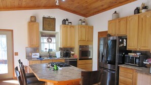 Une cuisine avec des meubles en bois, un îlot central doté de chaises, des appareils électroménagers en acier inoxydable et une fenêtre décorée d’une guirlande.