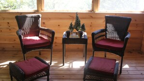 Deux fauteuils en osier avec des coussins rouges, une petite table sur laquelle est posée une plante en pot, et l’intérieur d’un chalet en bois doté de grandes fenêtres.