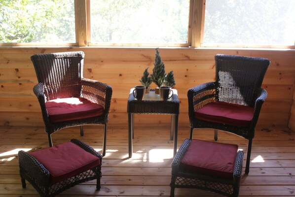 Two wicker chairs with red cushions, a small table with a potted plant, and a wooden cabin interior with large windows.