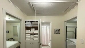 A room with a bed, a closet with folded towels, a bathroom with a shower curtain, and a door leading to another room. - Mid Century Modern Retreat in North Augusta (North Augusta)