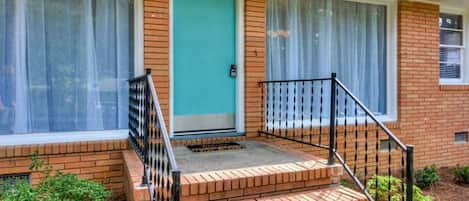 A two-story brick house with a front porch, a blue door, and a small set of steps leading to the entrance.