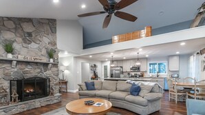 A spacious living area with a stone fireplace, a ceiling fan, and a kitchen in the background.