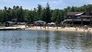 A lakeside beach with a dock, picnic tables, and a building with a stone facade.