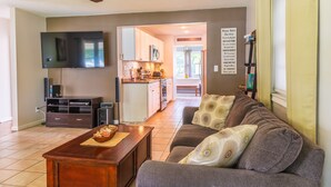 A living room with a sofa, a coffee table, and a TV. There is a kitchen in the background with cabinets and a microwave.