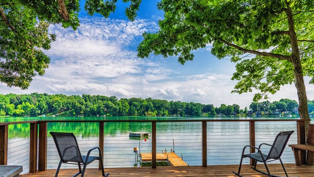 A wooden deck with two rocking chairs overlooking a lake with a dock and a small boat.