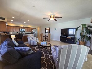 A living room with a sofa, armchairs, a coffee table, a TV, a ceiling fan, and a kitchen in the background.