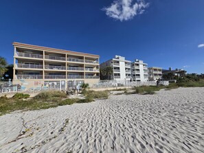 A beachfront hotel with multiple balconies, a sandy beach in the foreground, and a clear blue sky.