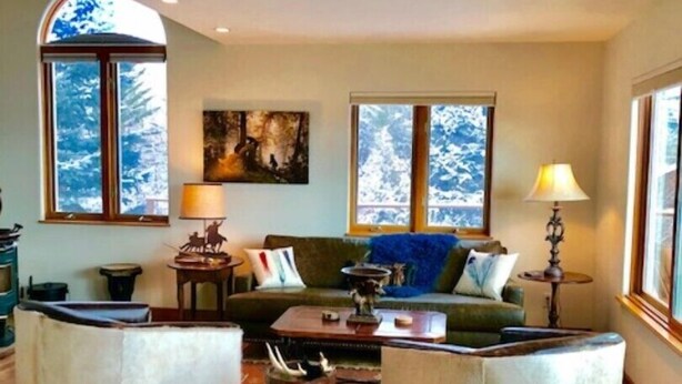 Un salon chaleureux avec un canapé, deux fauteuils, une table basse et une cheminée. Deux fenêtres offrent une vue sur des arbres couverts de neige.