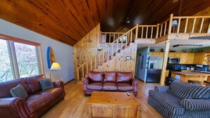 A spacious living area with wooden flooring, a brown leather sofa, a striped sofa, and a wooden coffee table. A staircase leads to an upper level with a balcony.