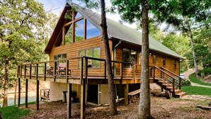 A wooden cabin with a deck, large windows, and a metal roof, surrounded by trees and a body of water.