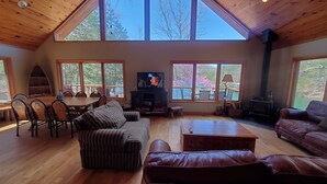 A spacious living room with a high wooden ceiling, large windows, a fireplace, and a television.