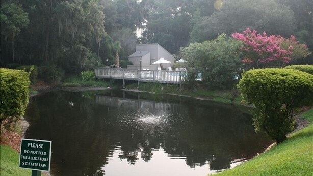 A pond with a sign prohibiting feeding alligators, surrounded by lush greenery and a house with a deck. - Newly Updated, Near Harbour Town 1st Floor 2 Bedroom 2Bath  Villa  (Hilton Head Island)