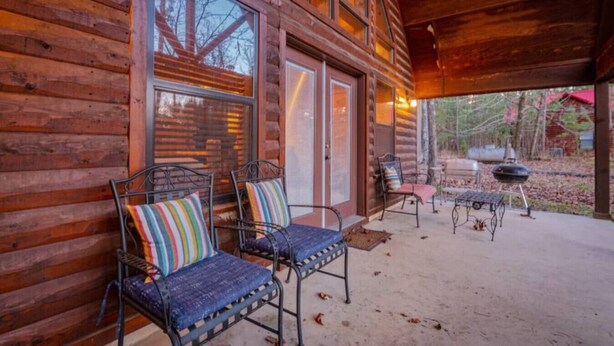 A wooden cabin with a covered porch, two metal chairs with colorful cushions, and a barbecue grill.