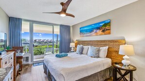 Une chambre d’hôtel avec un grand lit, un ventilateur de plafond, un tableau représentant une plage et une vue sur l’extérieur.