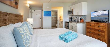 A bedroom with a large bed, a ceiling fan, a TV, and a kitchenette.