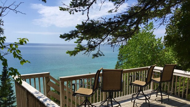 A wooden deck with chairs and a table overlooking a body of water. - Lake Michigan Home on Tunnel of Trees with Game Room and Views (Harbor Springs)