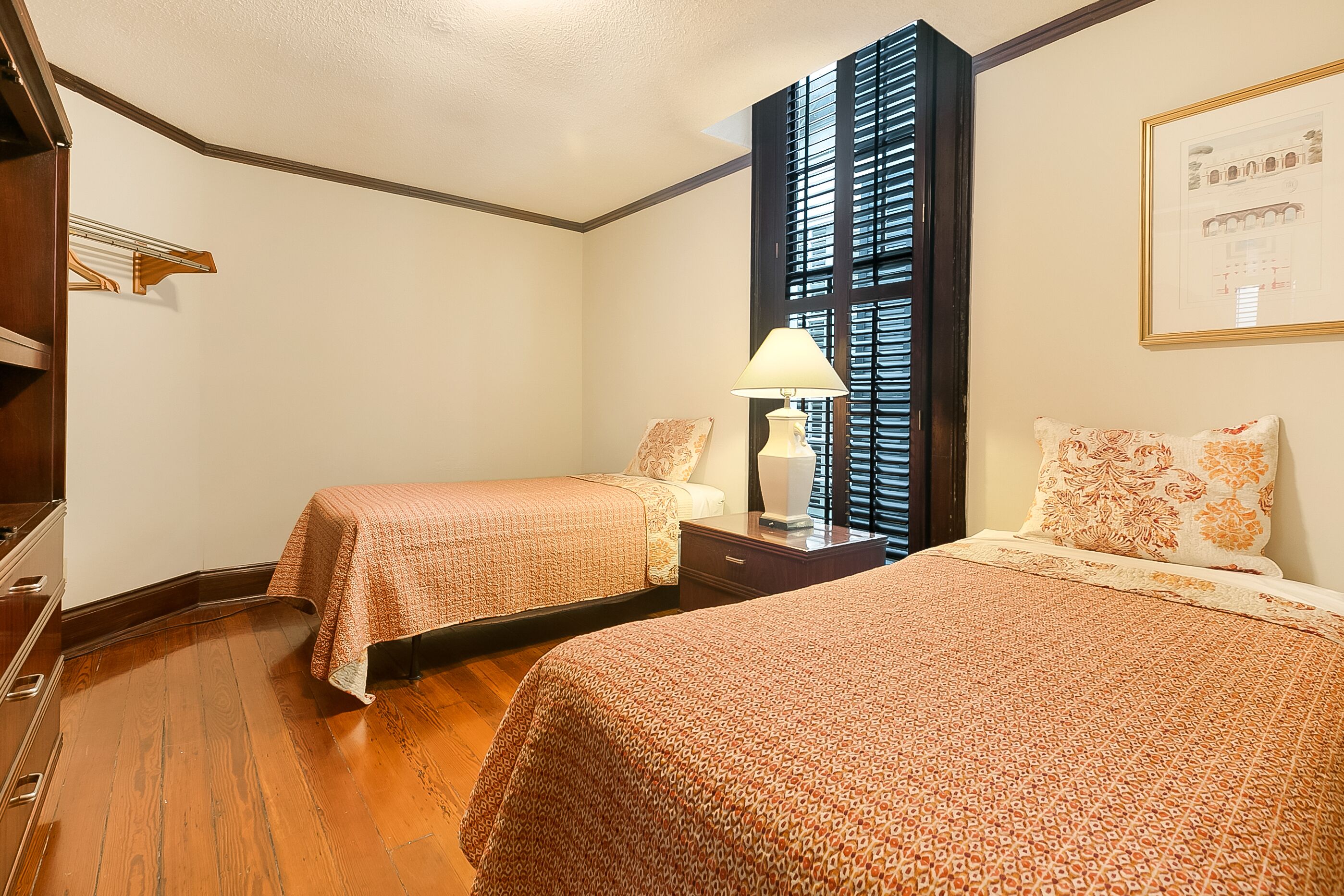 Habitación de hotel con dos camas, una mesita de noche con lámpara, un tocador de madera y una ventana con persianas.