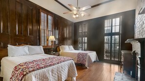 A room with two beds, wooden flooring, a ceiling fan, and large windows with shutters.