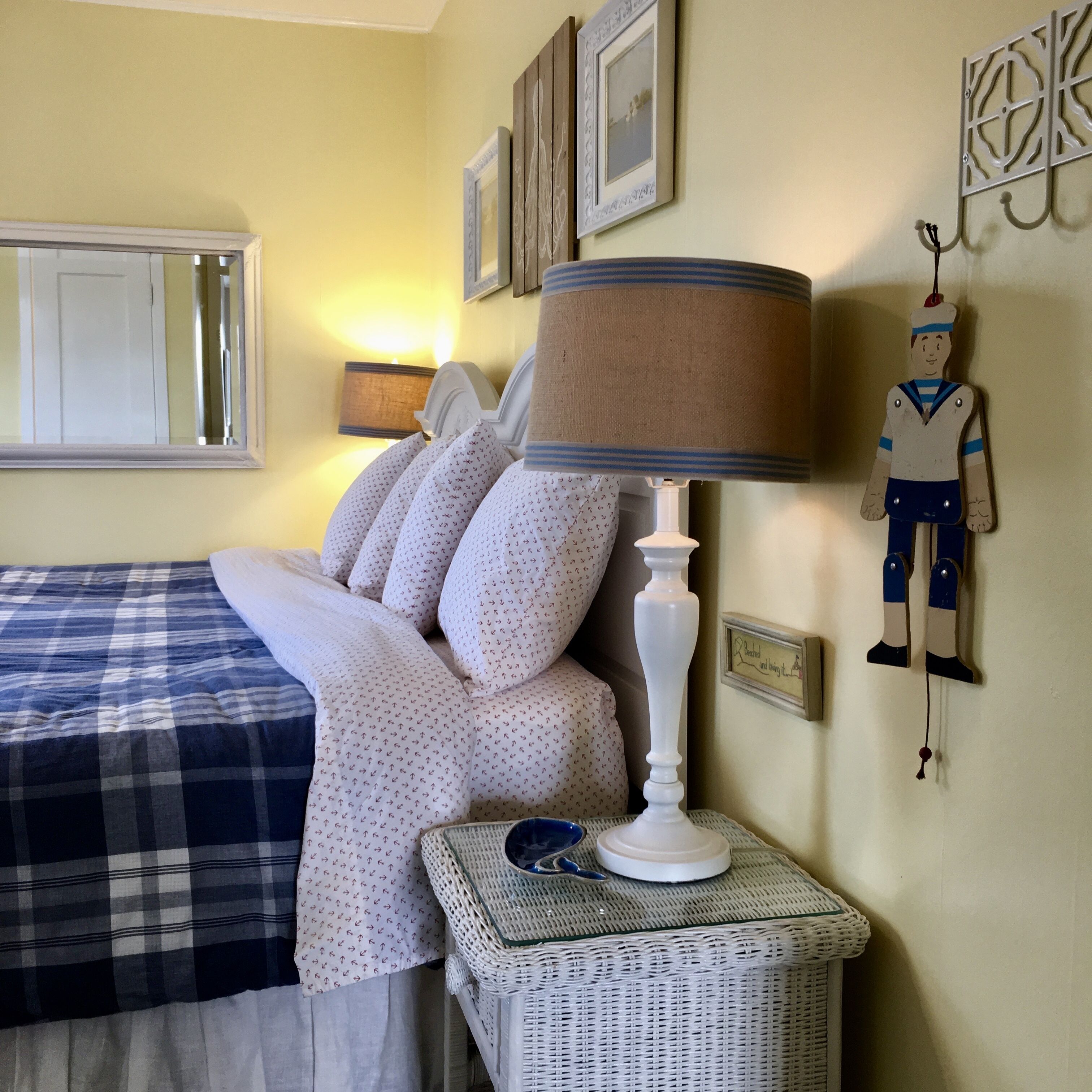 A neatly made bed with a blue and white checkered blanket, a white lamp with a beige shade, and a small wicker side table with a blue bowl.