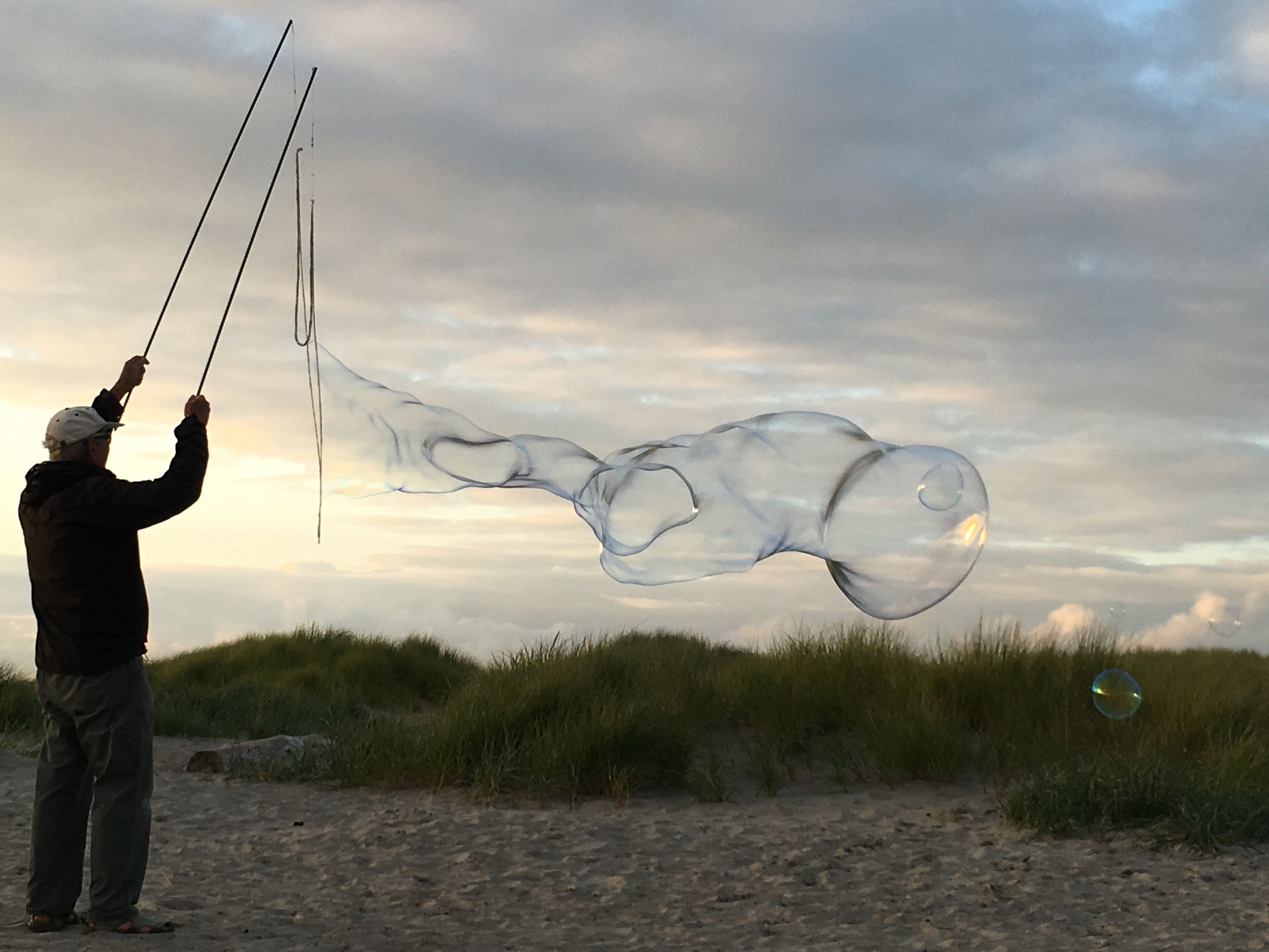 Eine Person lässt am Strand einen langen, durchsichtigen Drachen steigen, Dünen sind im Hintergrund zu sehen.
