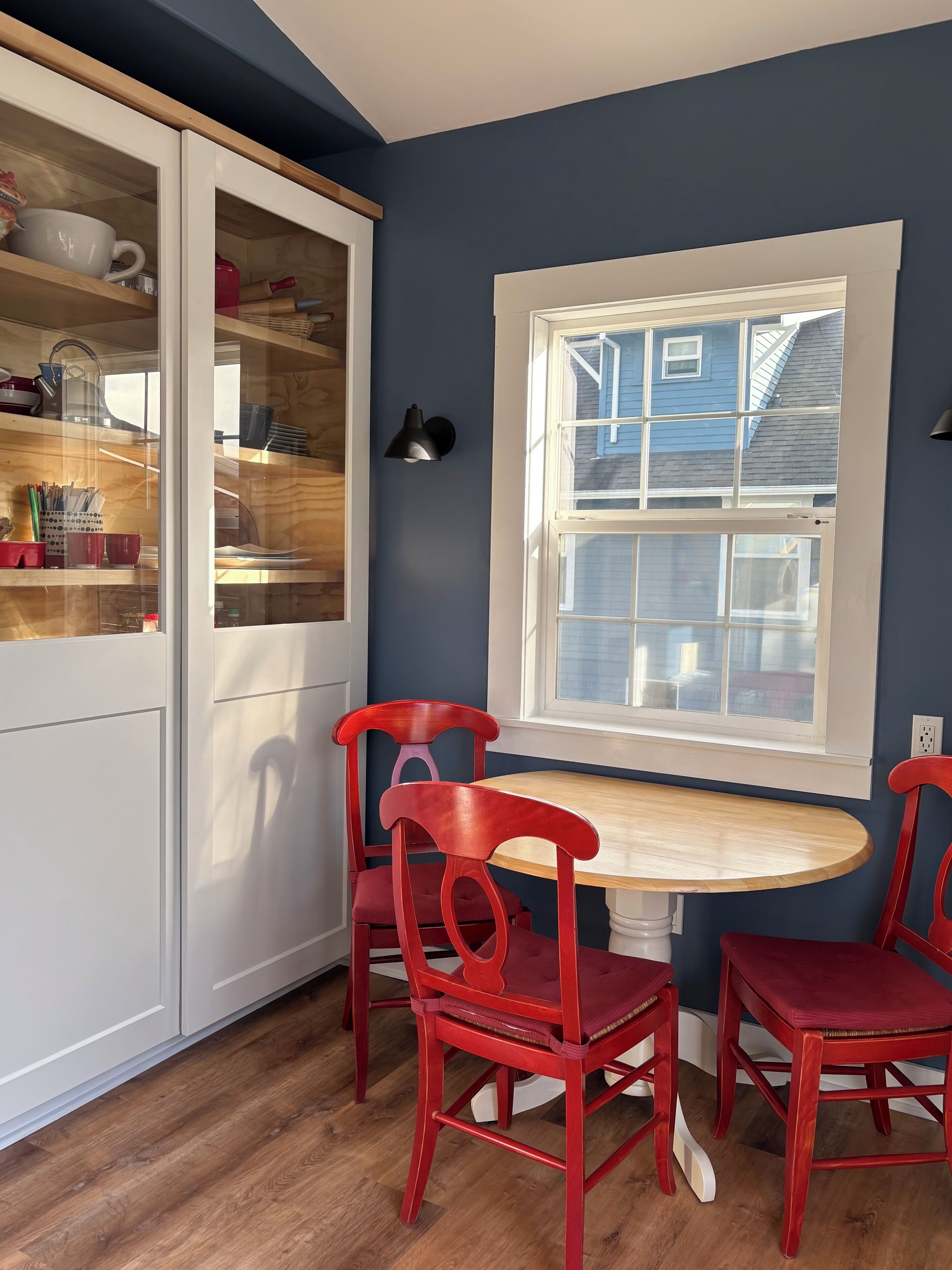 A dining area with a round wooden table, two red chairs, a white cabinet with glass doors, and a window with a view of a blue house.