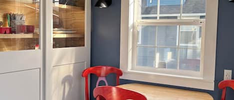 A dining area with a round wooden table, two red chairs, a white cabinet with glass doors, and a window with a view of a blue house.