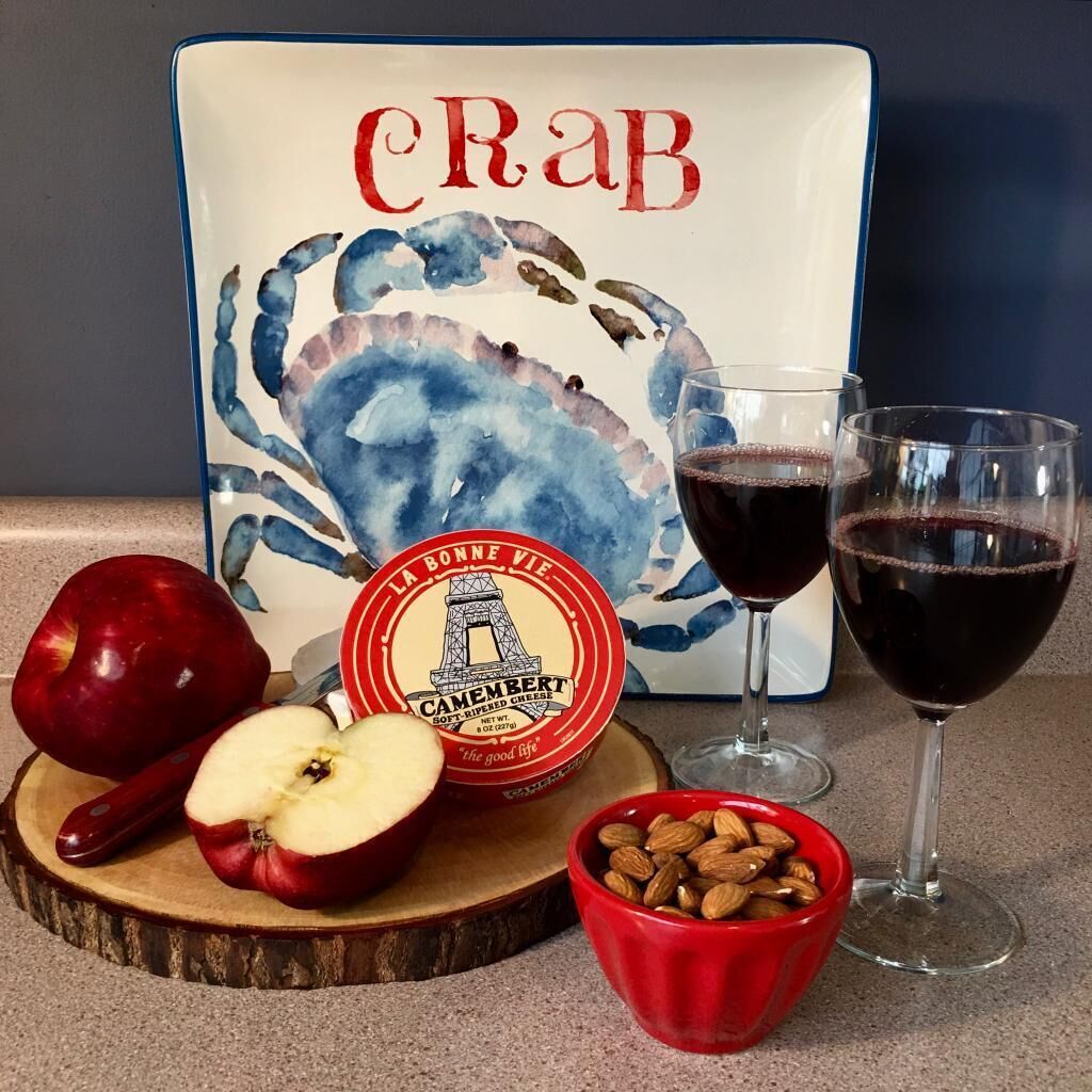 A table with a plate of food, two glasses of red wine, a half apple, a bowl of almonds, and a decorative plate with a crab illustration and text.