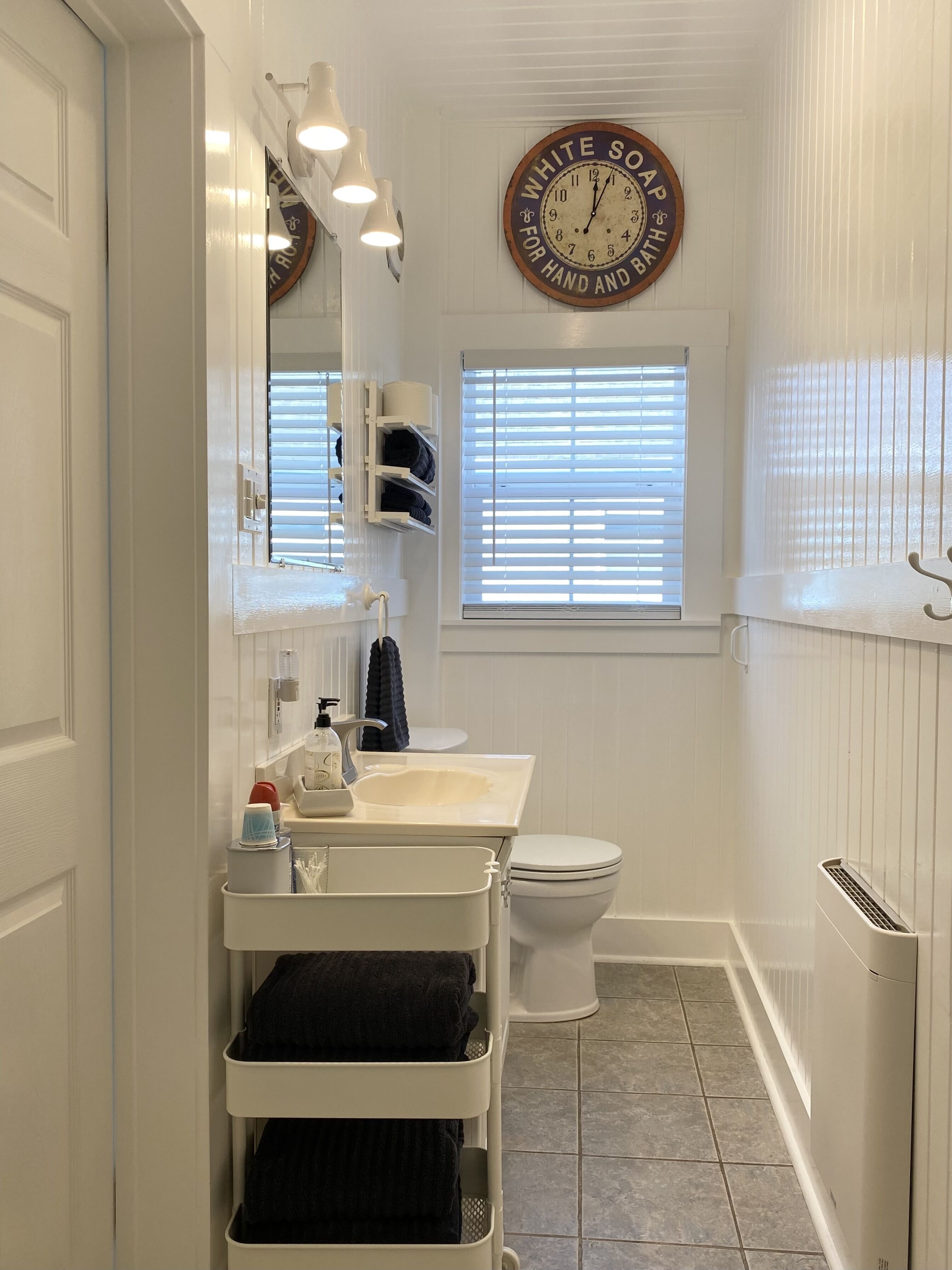 A bathroom with a white sink, a toilet, a towel rack, a window with blinds, and a wall clock.