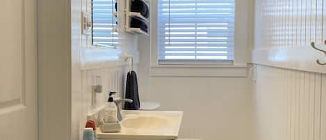 A bathroom with a white sink, a toilet, a towel rack, a window with blinds, and a wall clock.