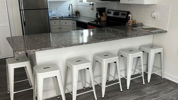 A modern kitchen with white cabinets, stainless steel appliances, and a granite countertop. There are four white stools and a stainless steel refrigerator.
