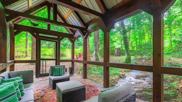 A covered outdoor seating area with wicker furniture and a colorful rug, overlooking a forested area with a stream. - Babbling Brook - 10 Minutes from Downtown Blue Ridge (Blue Ridge)