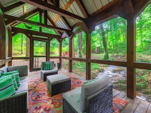 A covered outdoor seating area with wicker furniture and a colorful rug, overlooking a forested area with a stream. - Babbling Brook - 10 Minutes from Downtown Blue Ridge (Blue Ridge)