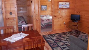 A wooden cabin interior with a dining table, chairs, a bed with a quilt, a television on a stand, and a framed picture on the wall.
