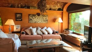 Ein gemütliches Zimmer mit Backsteinwand, einer Couch mit Kissen, einem Couchtisch, einer Lampe, einem Fernseher und einem Fenster mit Blick ins Grüne.