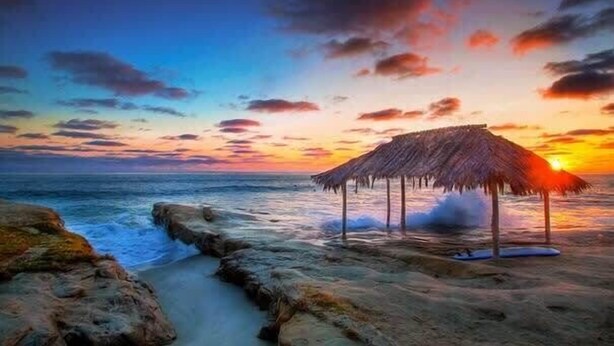 Una playa con una estructura de techo de paja, costa rocosa y olas del océano.