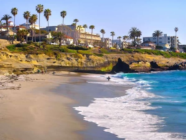 A coastal beach with rocky cliffs, palm trees, and buildings in the background.