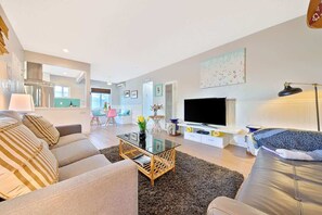 A modern living room with a sofa, coffee table, TV, and a dining area in the background.