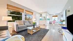 Una sala moderna con sofá, mesa de centro y televisor. Se observa una cocina al fondo.