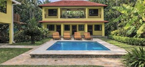 A two-story yellow building with a swimming pool and wooden lounge chairs in the front.