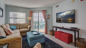 A living room with a beige sofa, a blue ottoman, a red sofa, a TV mounted on the wall, and a balcony with red and white striped curtains.