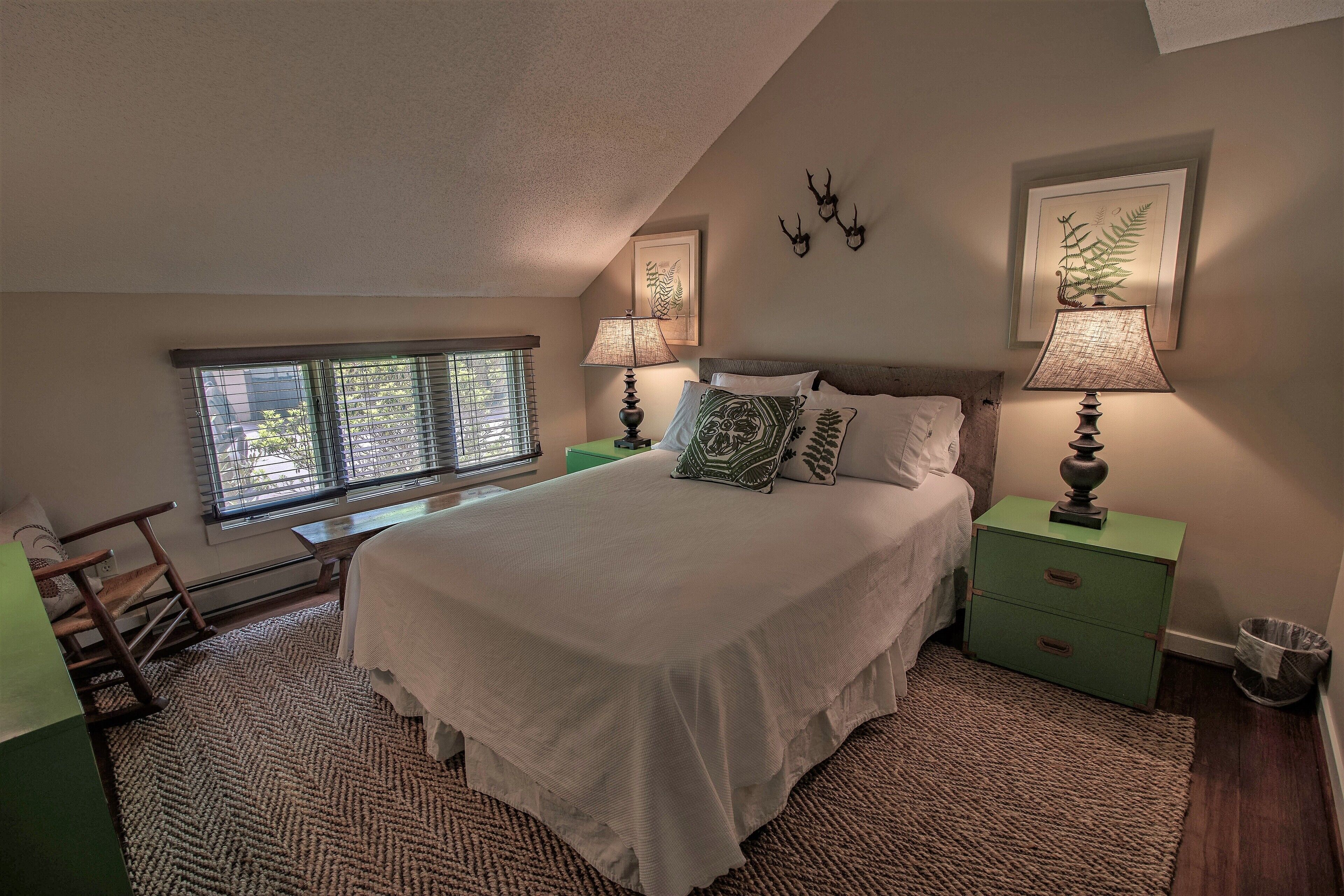 A bedroom with a large bed, two bedside tables with lamps, a window with blinds, a chair, and a green nightstand.
